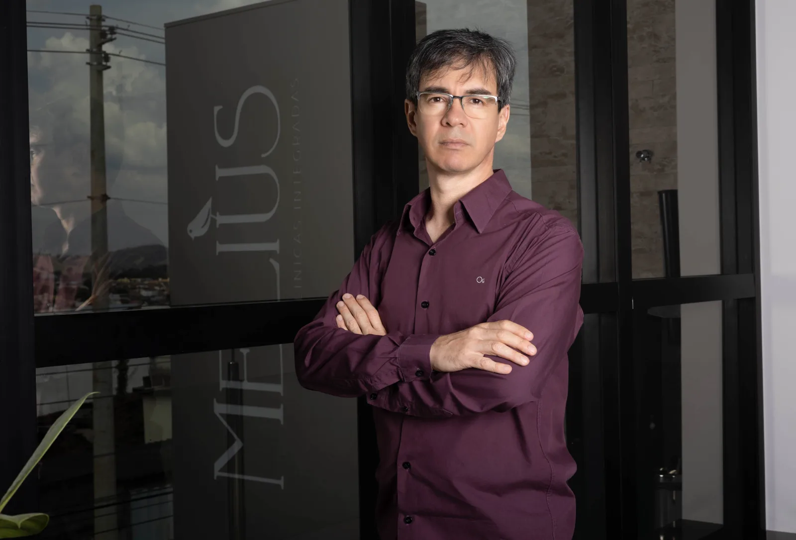 Dr. Alessandro de camisa vinho e braços cruzados, na entrada da Clínica Melius.